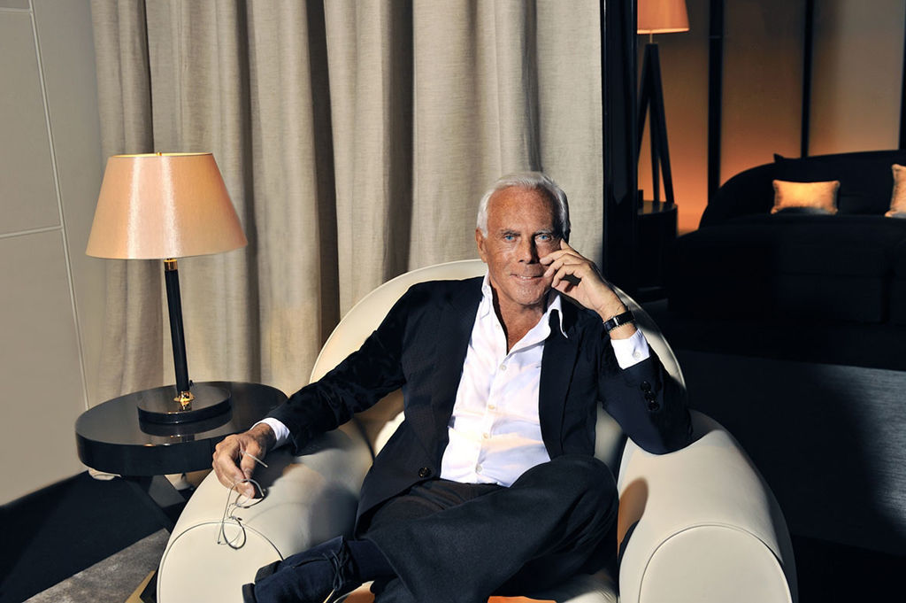 A portrait of Giorgio Armani, exuding elegance and sophistication as he sits in a stylish armchair, dressed in a classic black suit, with a thoughtful expression and a chic lamp in the background.