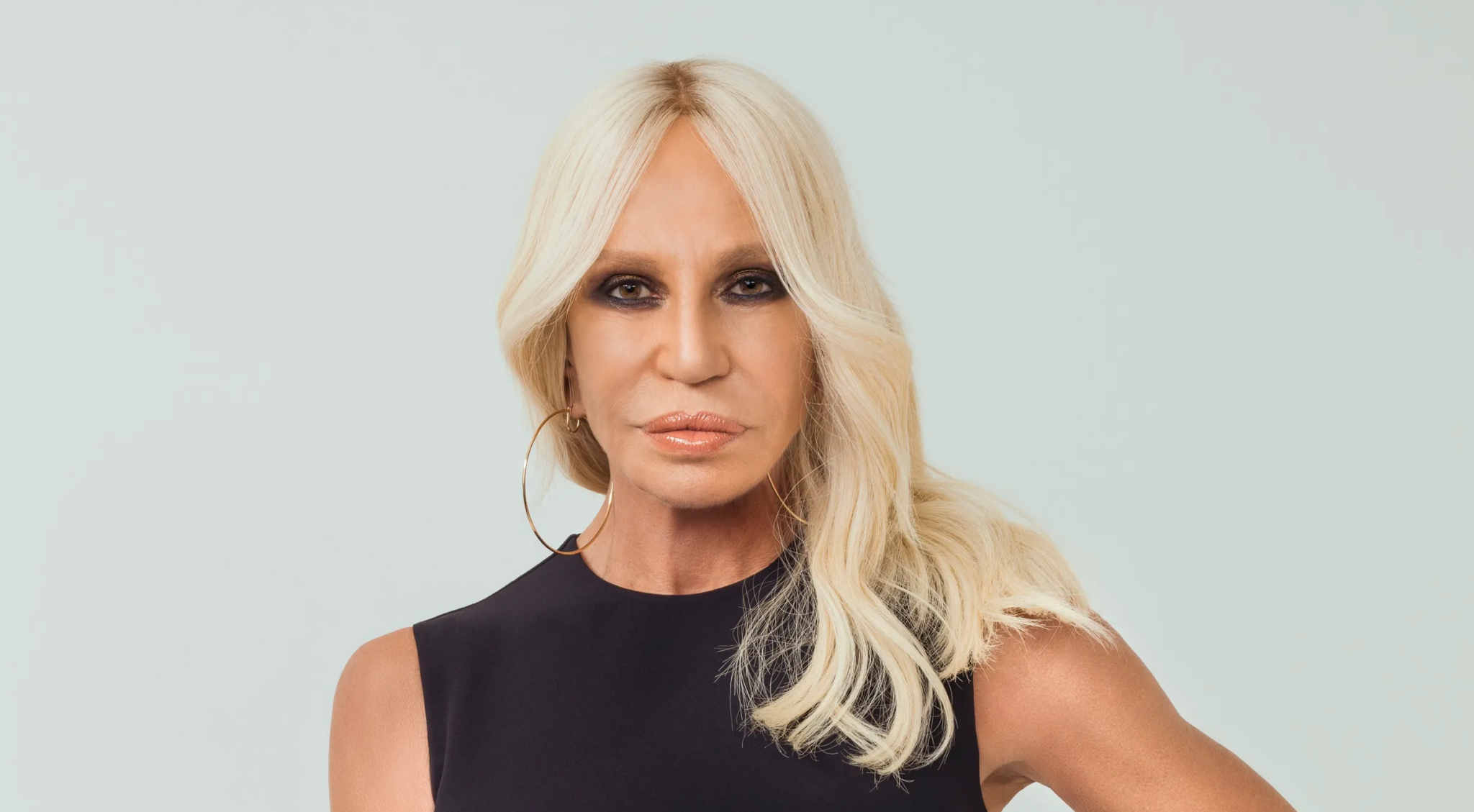 A striking portrait of Donatella Versace, showcasing her iconic style with long, wavy blonde hair, bold makeup, and statement hoop earrings, exuding confidence and elegance.