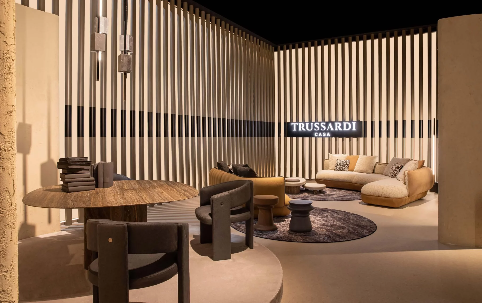 A modern living area by Trussardi Casa featuring a plush curved sofa, a stylish round table, and elegant seating, all set against a backdrop of vertical slats and warm lighting.