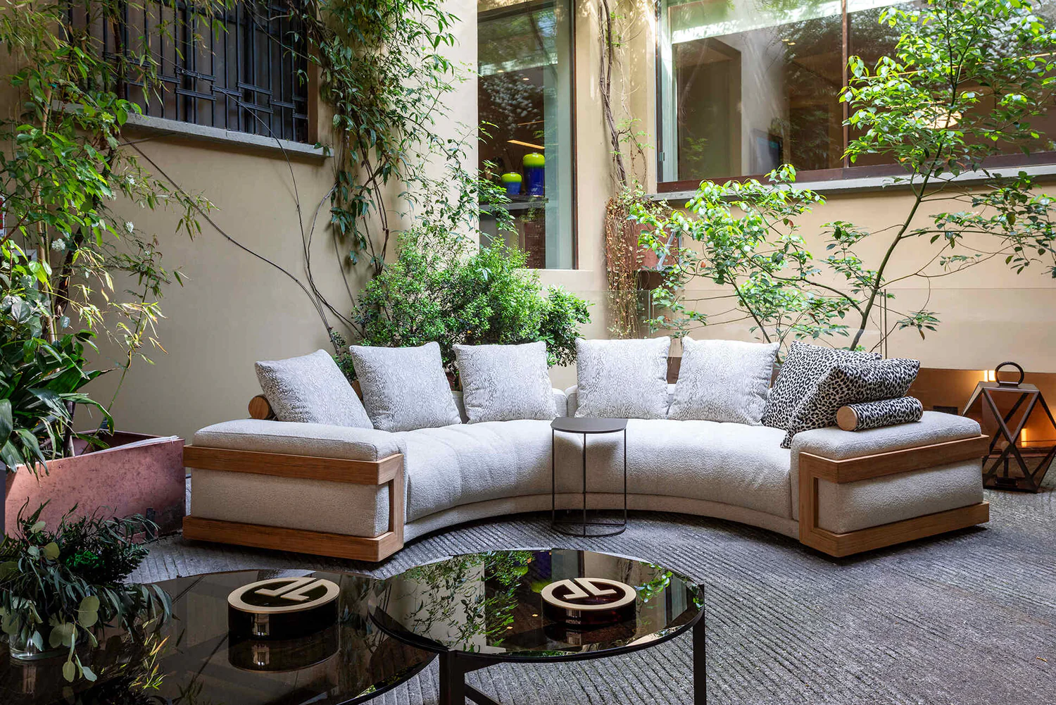 A luxurious outdoor seating area by Luxence Luxury Living featuring a curved, plush sofa with decorative cushions, complemented by greenery and a stylish side table, creating a serene and inviting atmosphere.